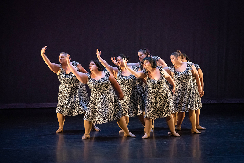 Seven dancers face front onstage and reach to their right side, all wearing black and white dresses