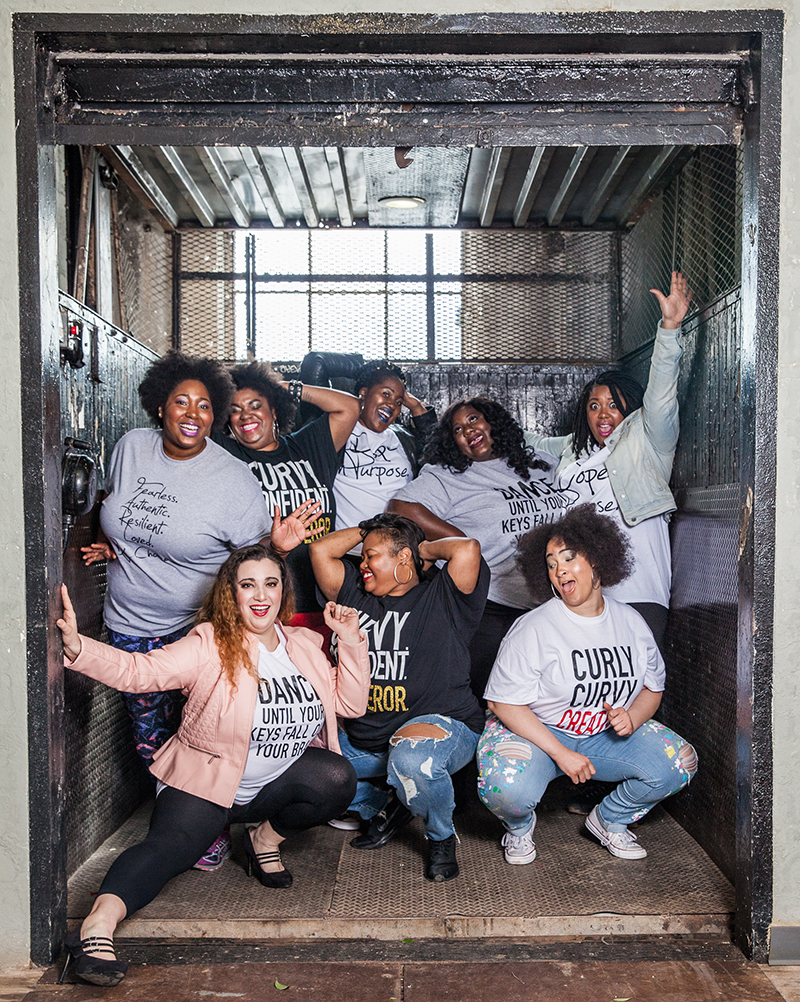 Eight fat women in two rows pose and smile, all wearing fat positive shirts.
