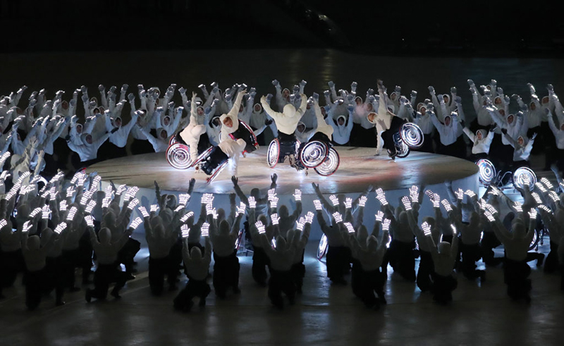 Five dancers in wheelchairs are on an elevated stage in a circle of hundreds of other dancers around them with their hands up.