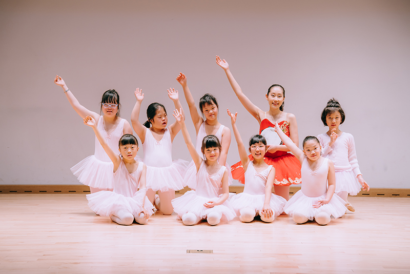 Eight young girls in pink tutus pose with their arms in the air with their teacher.