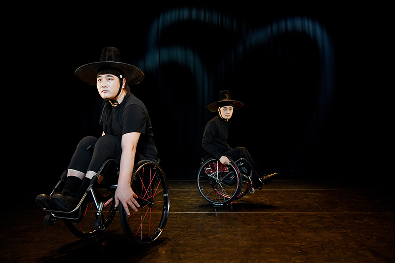 Two dancers are onstage leaning back in their wheelchairs wearing all black and black wide brimmed hats.