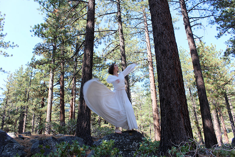 Natalie is mid-movement wearing a long white dress and dancing oudoors among big pine trees.