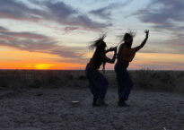 Two dancers dance in silhouette against a sunset in the desert wearing straw hats.
