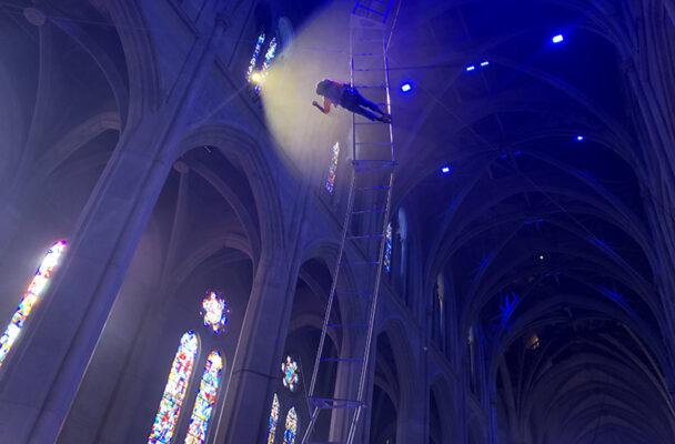 A dancer midway up a very tall ladder in Grace Cathdral leans toward a light.
