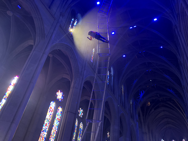A dancer midway up a very tall ladder in Grace Cathedral leans toward a light.