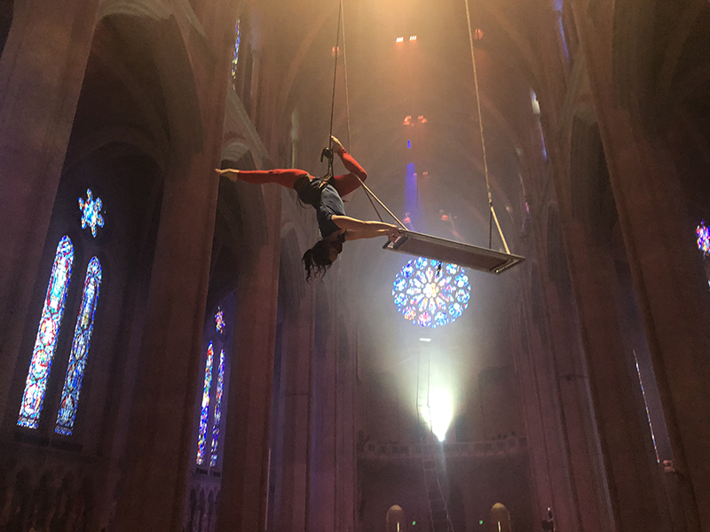 A dancer hangs upside down suspended from the ceiling of Grace Cathdral with her leg entwined around an enormous swing.