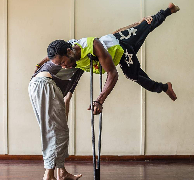 Tebandeke is suspended in middair with his legs in a V shape. He is supported by his crutch and another dancer.