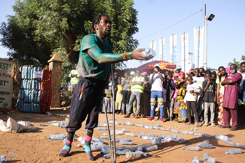 Tebandeke is outside on dirt surrounded by water bottles and holds one out in his hand. A crowd watches.