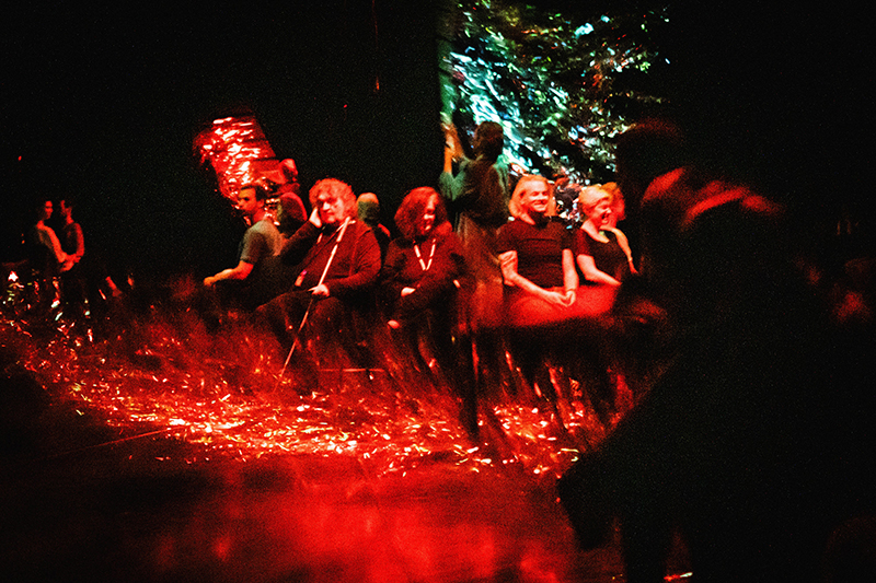 Five people sit onstage, one with a cane. The lighting is red, and a silvery set piece seems to swirl behind them. 