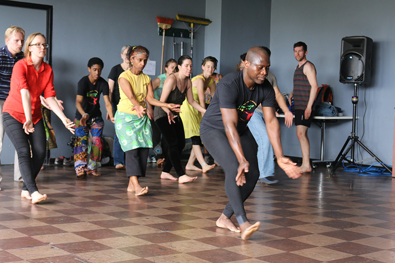 Etienne teaches a class to students, all have outstretched arm and one flexed foot.