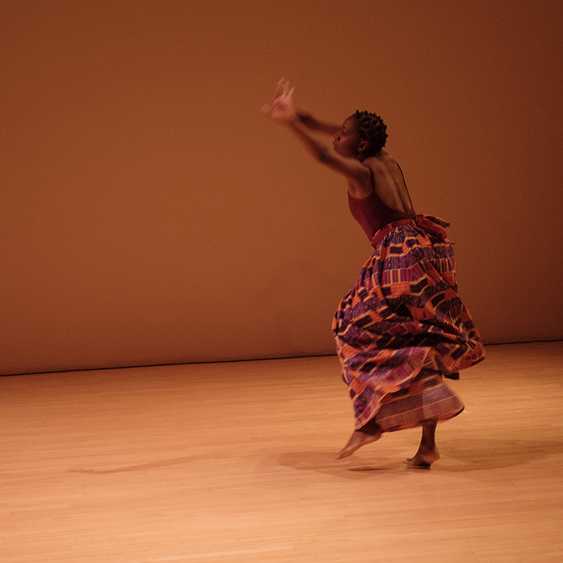 Rujeko on a wooden floor performing with her back to the audience and both arms raised. She is wearing a long skirt.