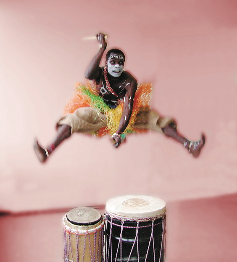 A dancer wearing white face paint jumps high above the drums in a straddle.