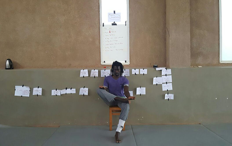 Momar sits in a chair on a sidewalk. Pieces of paper are taped to the wall behind him.