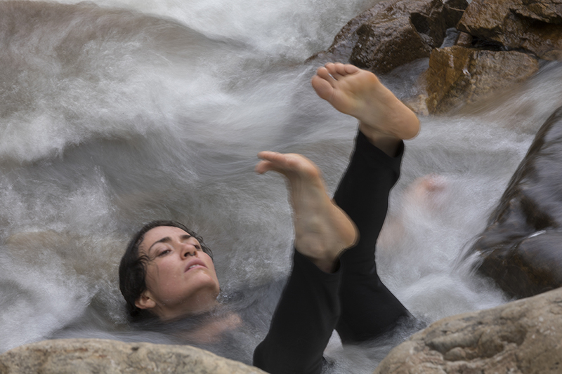 A person sits in a gushing stream with only their head and their legs and feet out of the water.