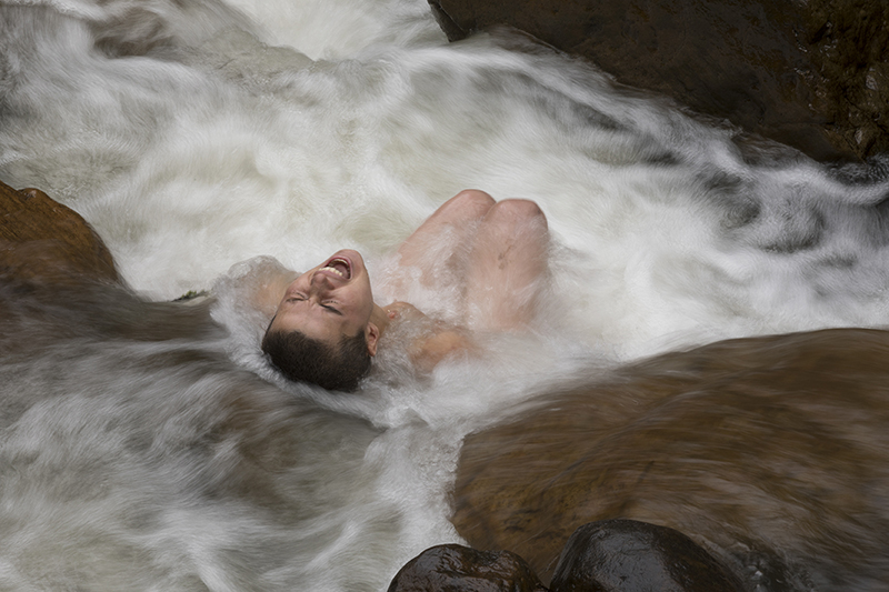 A naked person sits in a gushing stream with thei eyes closed and mouth open.
