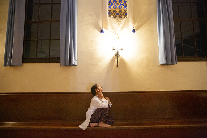 A dancer sits on a wooden pew looking up at a light.