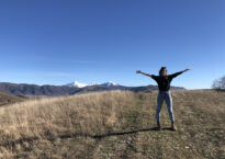 Giulia standing on a hill with arms wide open and mountains in the background.