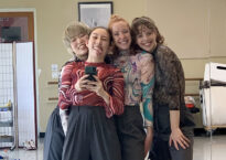 Four dancers bunched together smile into a mirror and take a selfie.