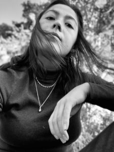 A closeup portrait of Feliticas in black and white wearing a turtleneck and a necklace with branches behind her.