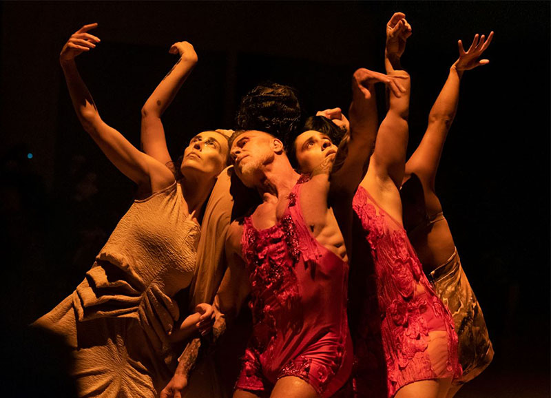 Seven humans in red and beige, feathered and pleated costumes lean backward toward each other, arms undulating like fleshy petals, and their upturned faces are bathed in a shaft of warm light. 