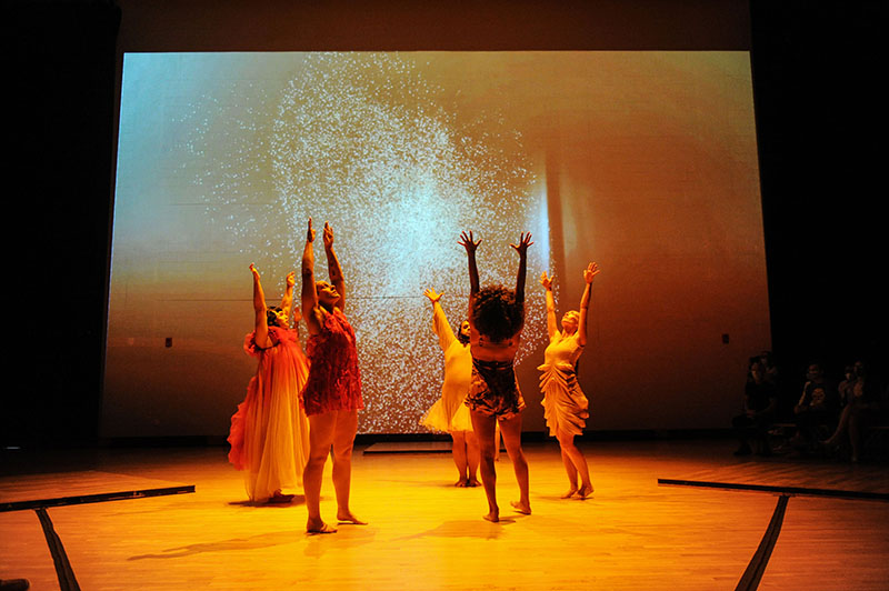 Seven humans in red and beige, feathered and pleated costumes stand in the center of a warm glowing space with their arms reaching high above; a projection of glowing, fragmented particles upstage.