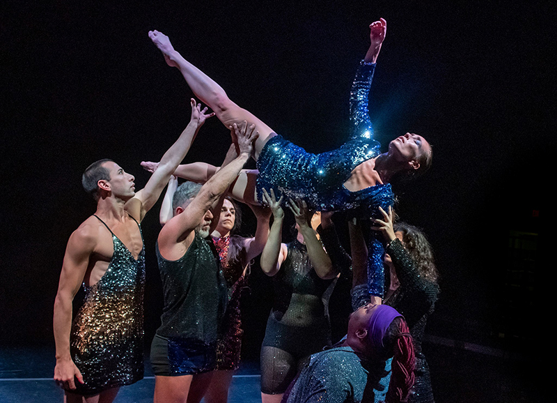 One shimmering human is held aloft by six others, legs straight and sharp, arm wearily reaching toward light, all wearing sequin costumes of gold, silver, black, blue, and purple. 