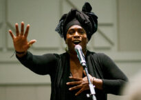 Soyinka is wearing all black with a black head wrap. She stands at a microphone with one hand on her heart and one hand outstretched.