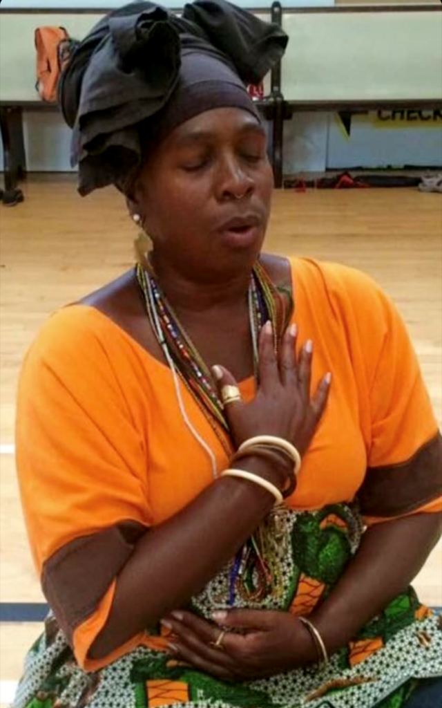 Soyinka sits with eyes closed and hands on her heart and stomach. She is wearing an orange shirt and a black head wrap.