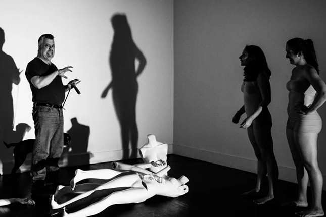 Joe gives direction to two dancers. Some dummies are lying on the floor. The photo is in black and white with stark shadows.