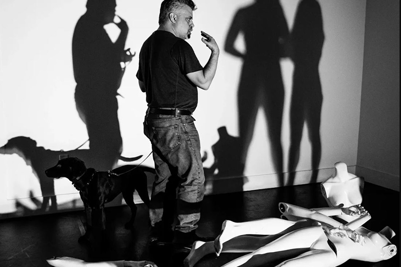 Joe and a dog stand over two dummies on the floor. The shadows of two dancers listening to Joe are cast on the wall behind him.