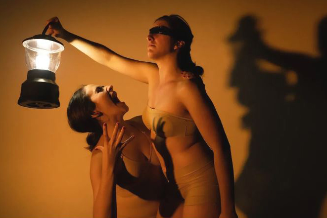 Two dancers in nude leotards are close together. One is blindfolded and holds a lantern. The other makes a face up at the first dancer.
