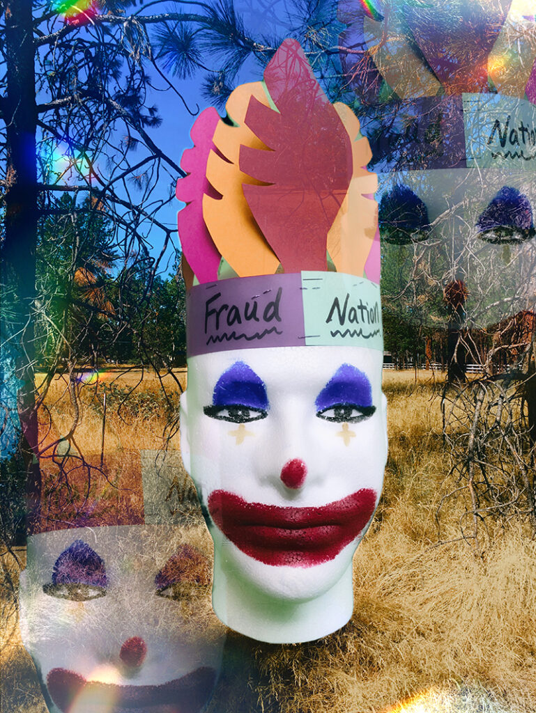 A clown mask with a paper feather headband reading "fraud nation" set among dried grasses.