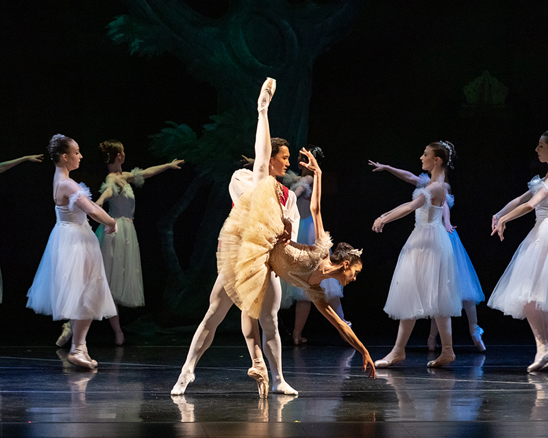 Michelle in a deep penché with a partner. She is wearing a yellow tutu and dancers in white tutus move behind her.