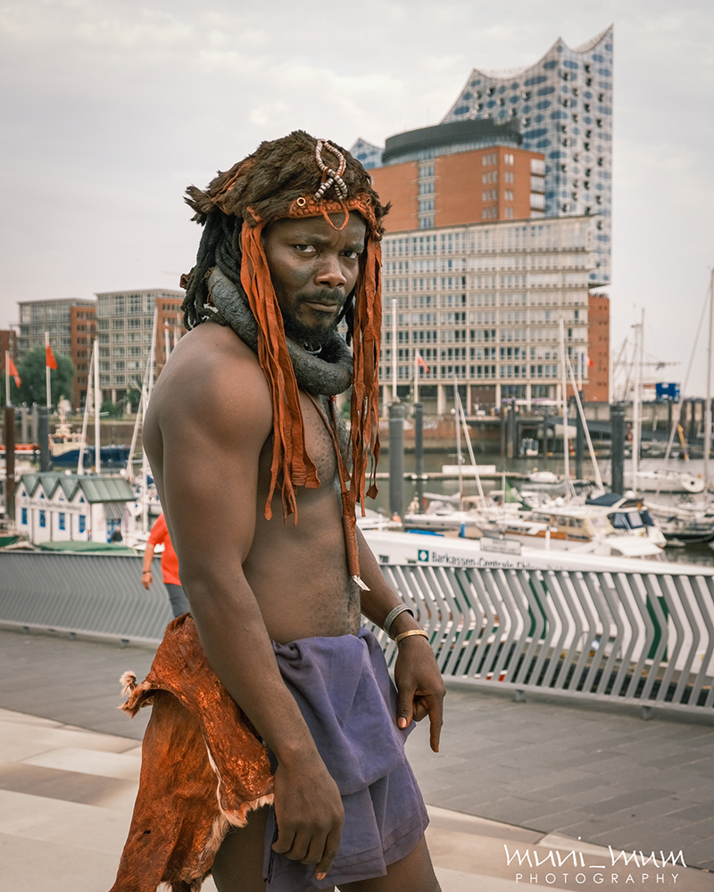 West stands in front of a modern cityscape wearing traditional Namibian garb.