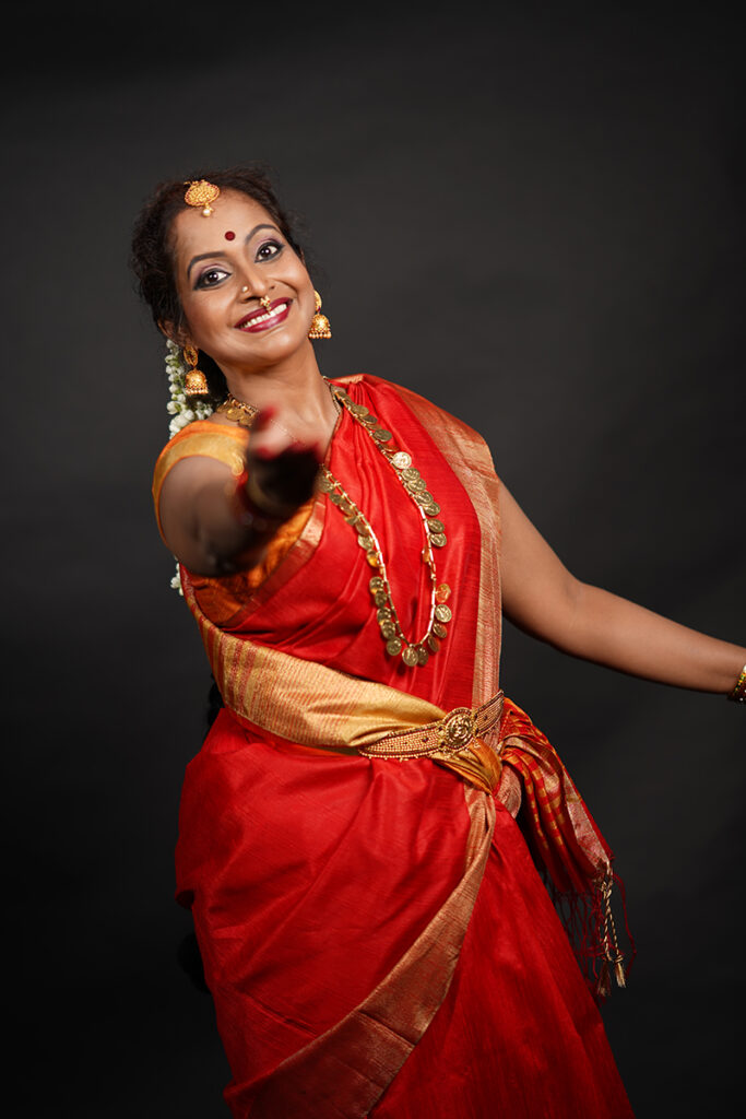 Yashoda smiles, wearing a red sari with gold jewelry. Her arm extends palm upward toward the camera. Her feet and other hand are out of the frame.