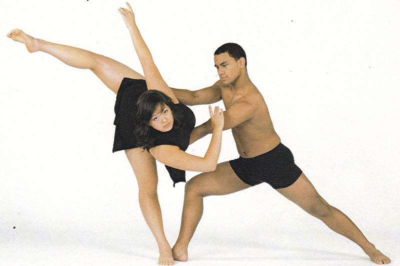 Aiano is being partnered by a young man holding their waist as they tilt away from him with their leg extended behind them.