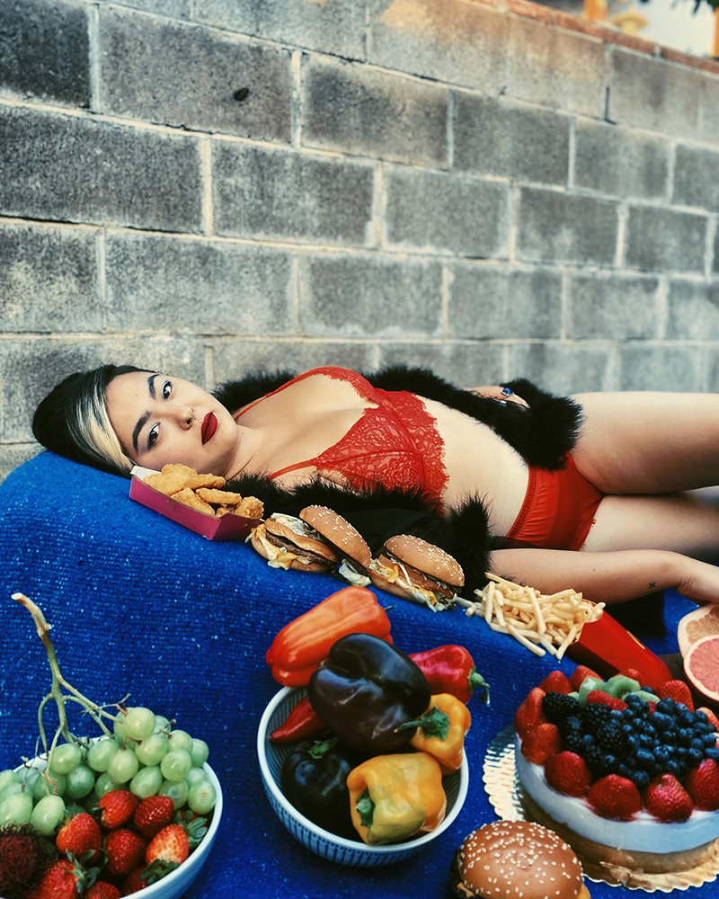 Aiano relaxes on a blue cushion wearing red lingerie. Plates of food adorn them.