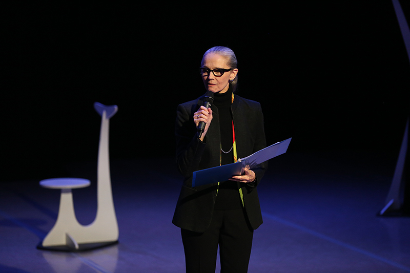 Janet stands and talks in a microphone on a dark lit stage. A podium is behind her and she holds a binder she's reading from.