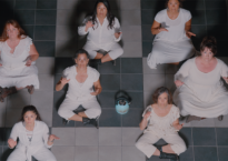Seven dancers wearing white sit crosslegged on a tiled floor and look up at the camera.