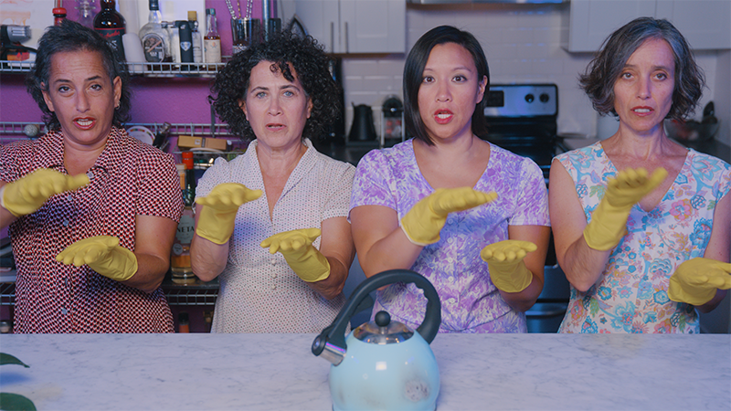 Four dancers sit at a counter top in front of a kettle with their hands in front of them. They are wearing big yellow kitchen gloves.