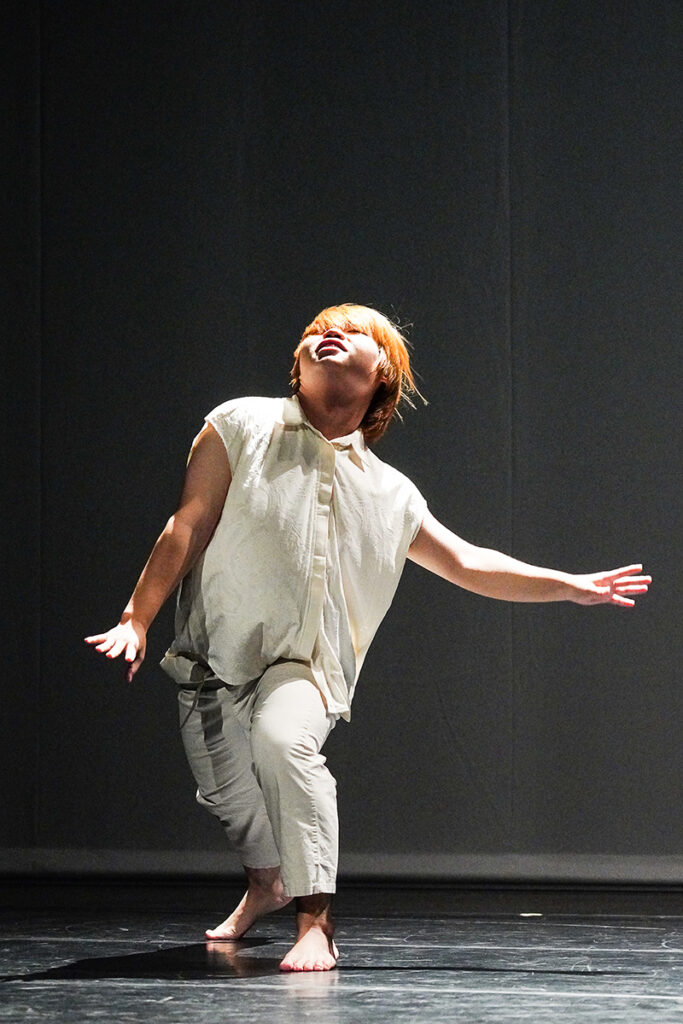 A dancer wearing white lunges onstage with hands gently extended to their sides. The dancer looks up.