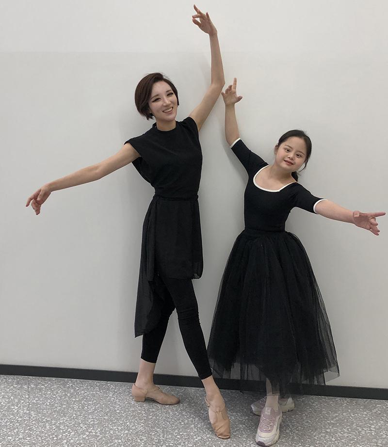 Two dancers pose in tendu against a wall. They are both wearing black.