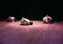 Three dancers hunched over in child's pose on stage with flower petals surrounding them.