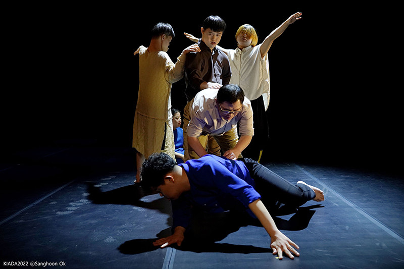Four dancers stand in a clump around one dancer lying on the floor. They are onstage against a black background.