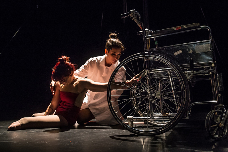 Erin and Vanessa sit onstage near an old-fashioned wheelchair. Erin reaches toward it and Vanessa reaches toward Erin. They are onstage. Erin wears red and Vanessa wears white.