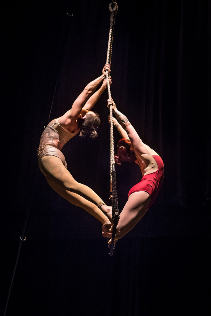 Vanessa and Erin are both holding onto a rope and leaning their torsos away from it while staying connected at the feet and in Erin's case the bottom of their legs. They are starkly lit onstage wearing leotards.