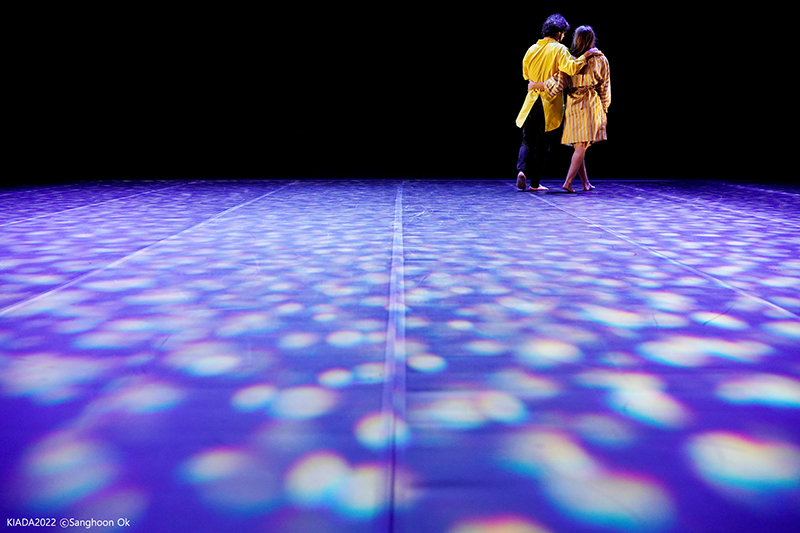 Helliot and Laura walk offstage arm in arm. The stage behind them is bathed in dark blue light.