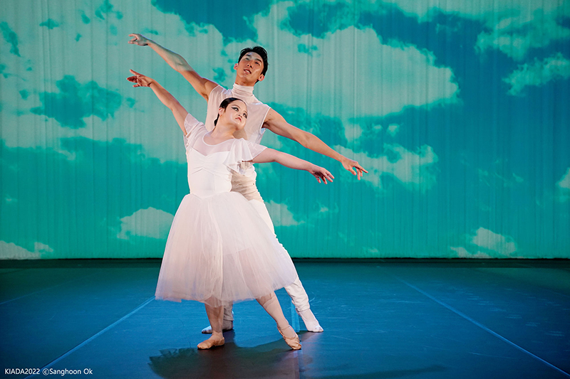 Two ballet dancers wearing white tendu to the side. The man is behind the woman. The backdrop is blue and green.