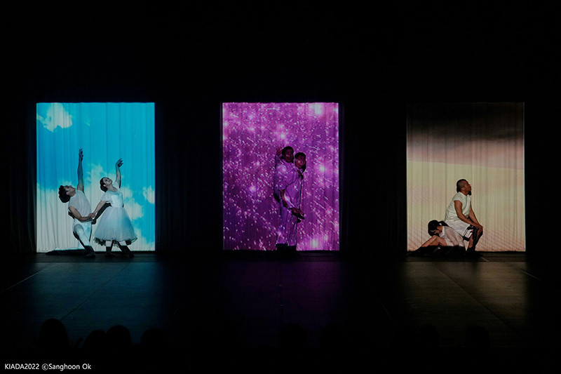 Six dancers pose onstage, two dancers each in three rectangles of light. One rectangle is blue, one is purple, and one is brown,