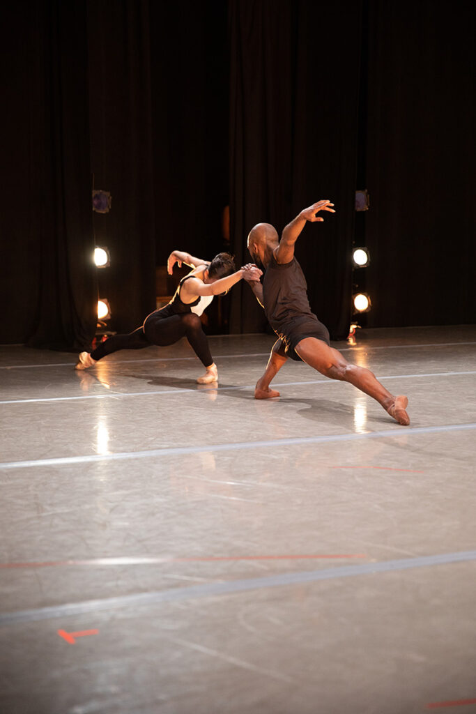 Two dancers onstage hold hands and lunge deeply toward each other.